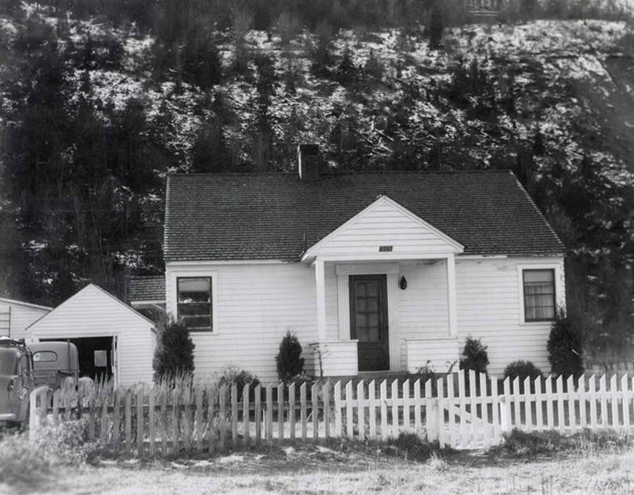 View of a house in Wallace, Idaho with a fence around it and cars parked in the driveway. A real estate view taken for Idaho First National Bank.