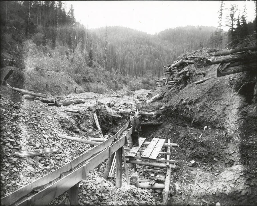 North side, Coeur d'Alene Mining District (Murray area).  A man stands next to the flume in the photograph.