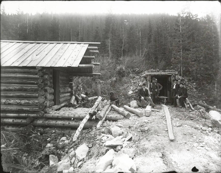 Men and a dog stand outside of the entrances to an unidentified mine and cabin.