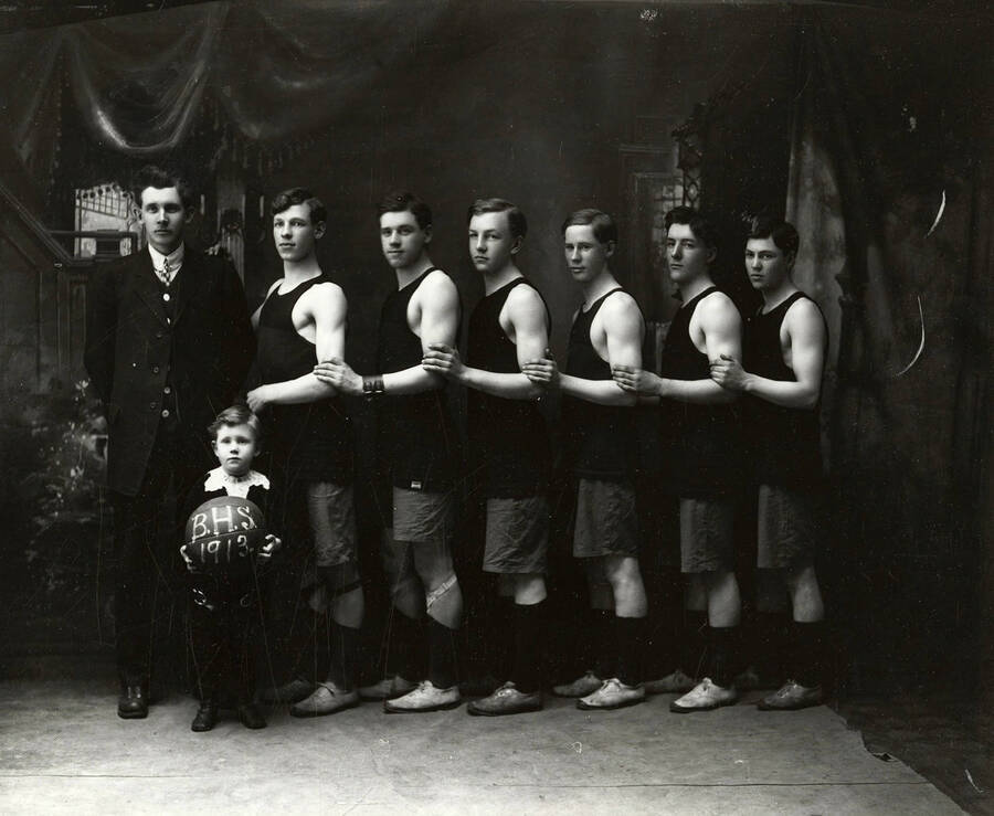 Group photo of the Burke boys' basketball team. Pictured are six players, one man, and a small boy holding a basketball that says "BHS 1913" on it.