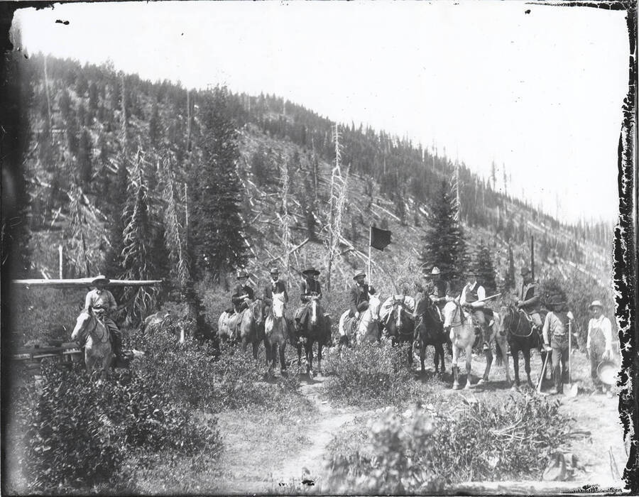 Men mounted on horseback. One man is carrying a flag, another a shovel and pick, and another a panning plate. Slight deterioration along edges of negative prior to photograph being printed.
