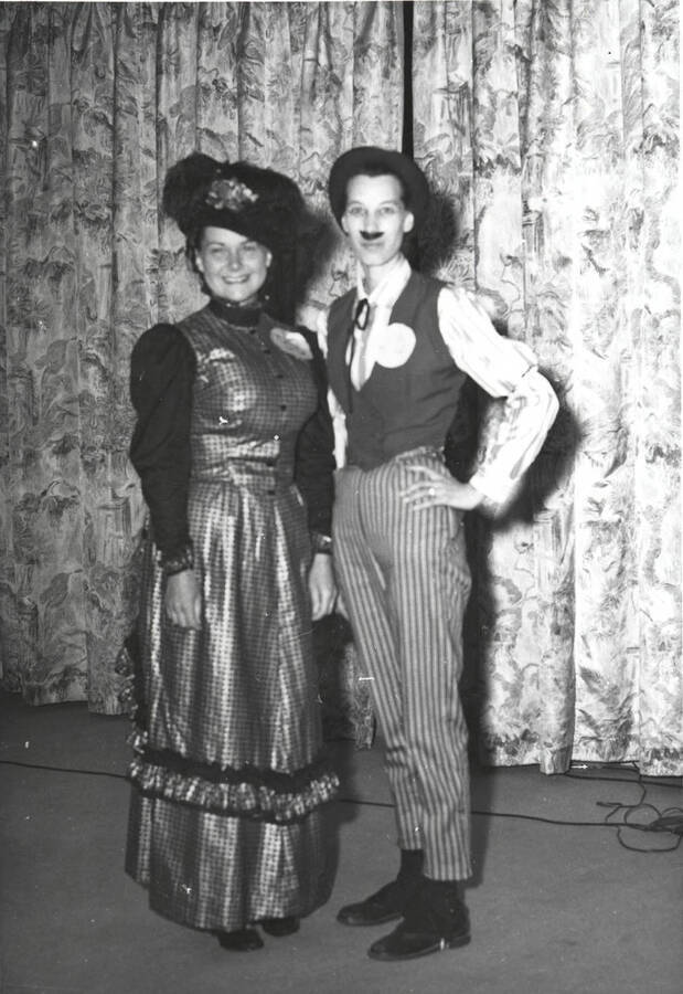 Formal portrait of two women in costume, one in a mens outfit and a false mustache, the other in a afternoon gown, at the North Idaho Press Jubilee.