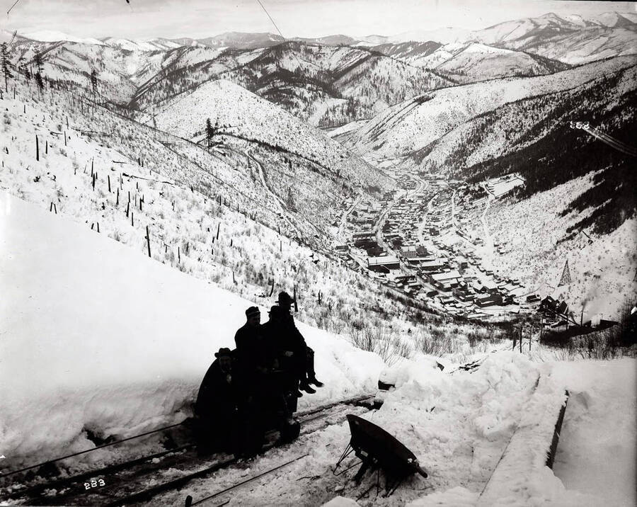 Winter view of Wardner, looking down from the Last Chance Mine. Four shaded men sit on an ore car.