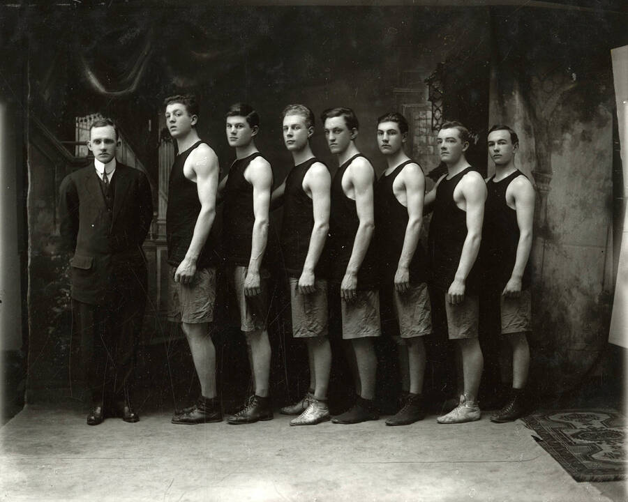 Group photo of the boys' basketball team of Wallace High School in Wallace, Idaho. Pictured are seven players and one man, all standing.
