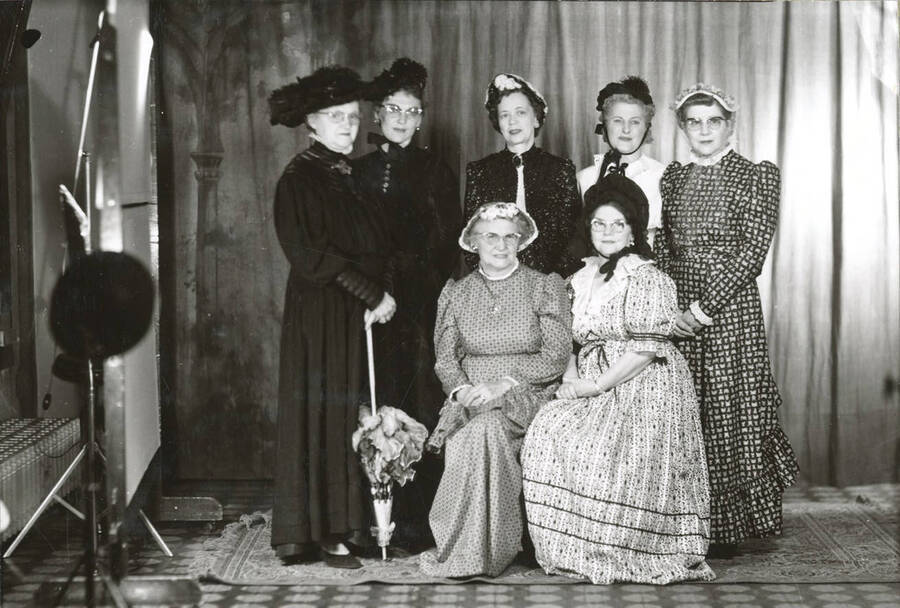 Formal portrait of a group of ladies in attendance at the North Idaho Press Jubilee. One of the stand lights is visible on the left hand side, as well as the edge of the backdrop and the radiator on the wall.