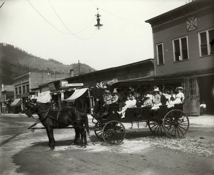 Group photo of the Sunday school group of the First Methodist Episcopal Church in Wallace, Idaho. The children are seen sitting on a wagon. The picture was taken on Cedar Street, at the end of 7th Street. The Wallace Fire Department and City Transfer can be seen in the background, along with other businesses.