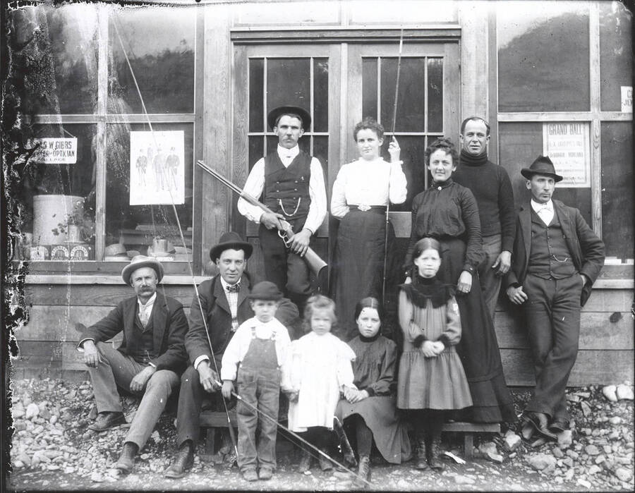 Men, women, and children, in front of a store in Wallace, Idaho. One man is holding a rifle while the children, women, and two other men hold fishing poles.
