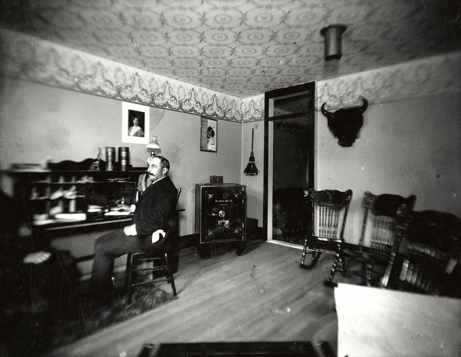 Herman J. Rossi sitting in a chair in an office. Behind are three rocking chairs, a safe, and a mounted bison's head.