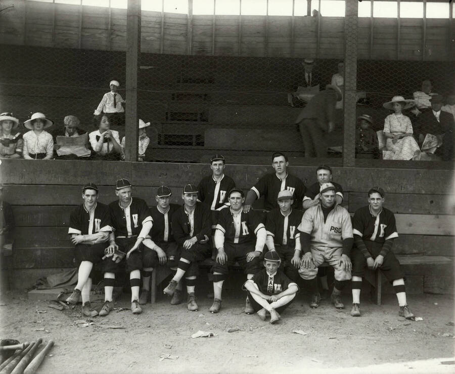 Group photo of the men's baseball team in Wallace, Idaho. Pictured is one coach, ten players, and spectators seated behind.