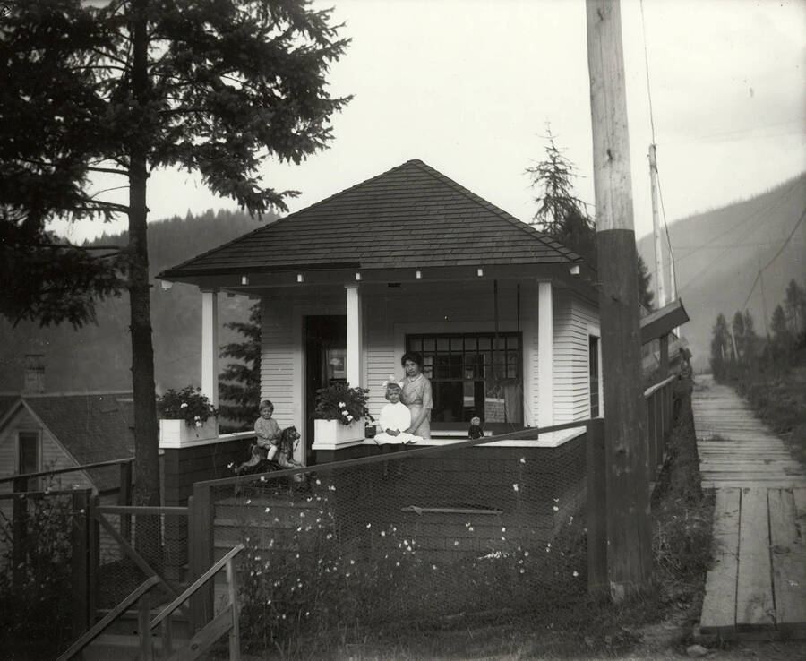 Photo of the Samuels family on the porch of a house. A woman stands with a little girl, while a boy sits on the rocking horse.