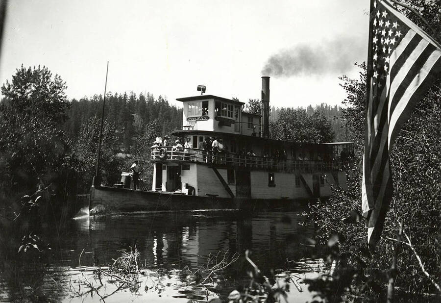 The steamboat "Colfax", which was built in 1902, on the water. People can be seen standing on the boats deck, and an American flag is in the foreground.