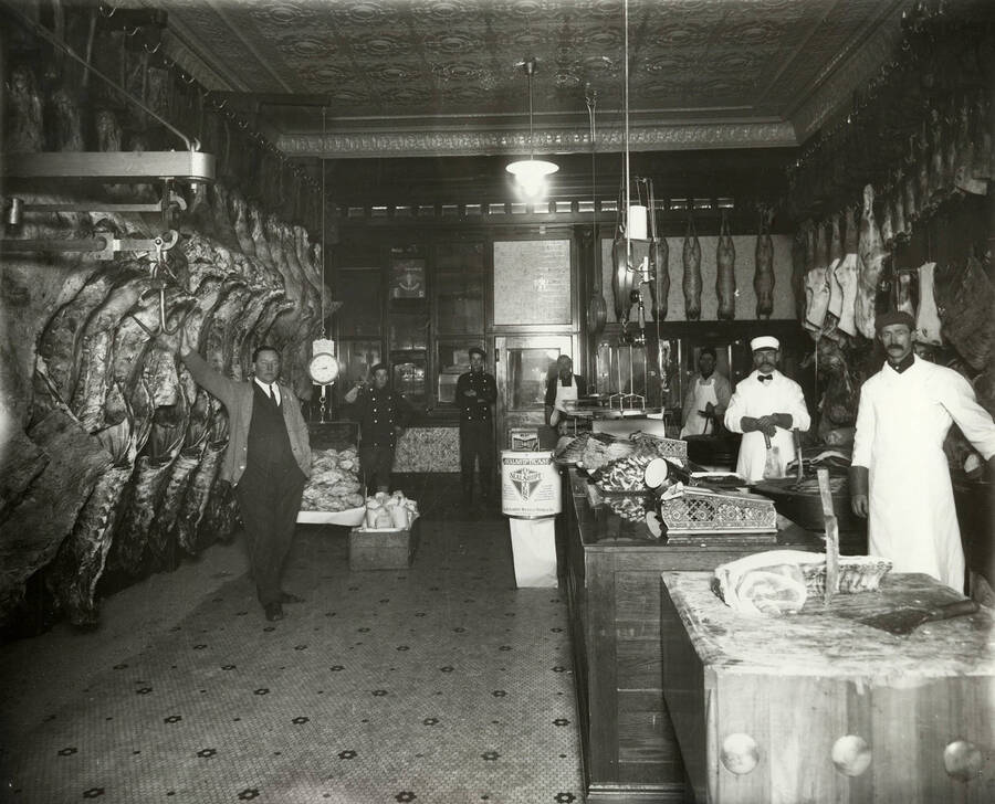 Interior view of Stevens Meat market in Wallace, Idaho. The picture shows large amounts of meat hanging, butcher blocks, scales, and eight men This market is located at 609 Bank Street and was owned by Fred A. Stevens.