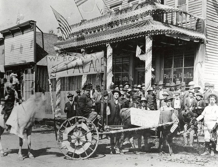 A cart decorated with a sign that reads, "We board at the Palace Hotel". The cart is parked in front of the Palace Hotel. 1. Panama Bill 2. Parley Thompson 3. Johnny Wiegele 4. Farmin Totten (Nova Scotia Carlson) 5. Howard Dodge 6. Frankie Harper 7. Robert Dodge 8. Joe Larenque (against post) 9. Godfrey Cole 10. Bert Murray.
