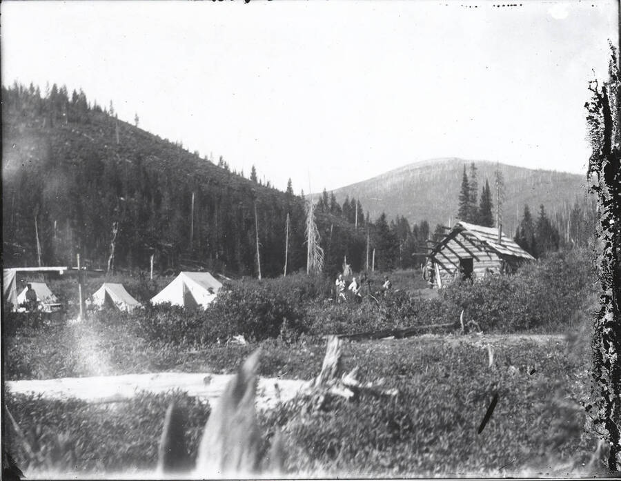 Men standing outside of a log cabin near tents and other man-made structures.