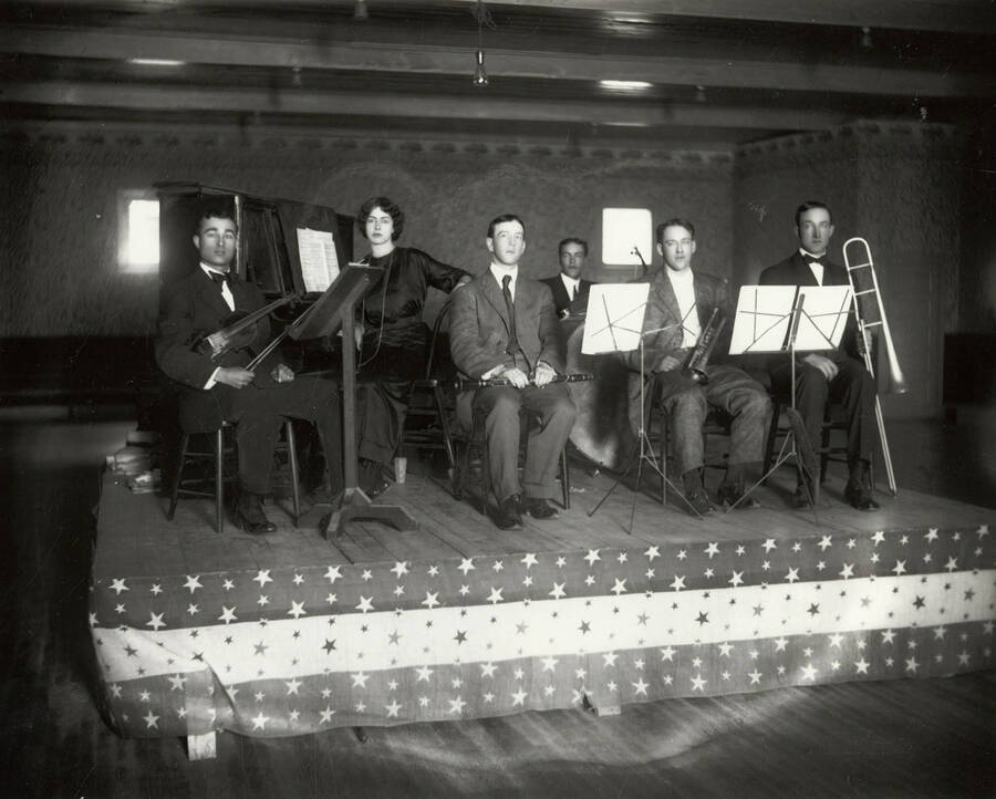 Group photo of an orchestra or dance band. The photo shows a six piece band, with one woman near the piano and five men seated and holding instruments. O.J. Lyons is part of this band.