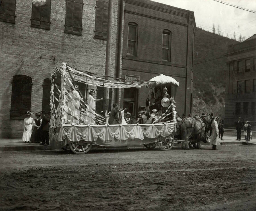 View of the Rebekah float. The wagon is decorated with a king, gauds, ladies, and children. The children are playing the horns. The float is shown on Hotel Street by the I.O.O.F. Hall in Wallace, Idaho.