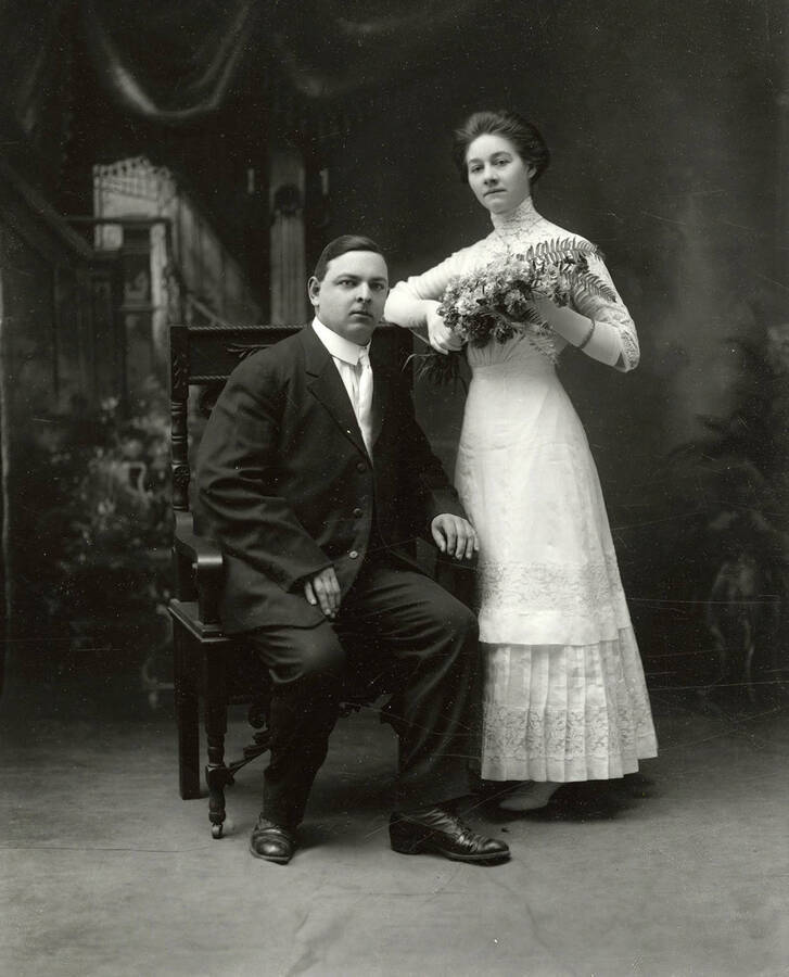 Photo of Mr. and Mrs. R.W. Bittell. Mr. Bittell is posed seated with Mrs. Bittell, who is standing and holding flowers.