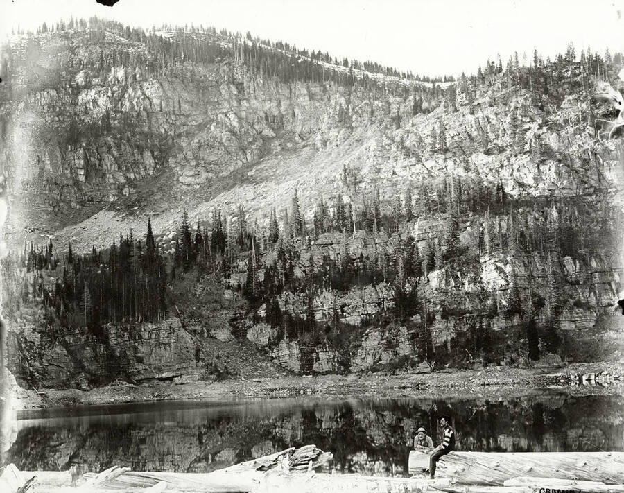 Two men sitting on the shore of Lower Stevens Lake near the Montana border in what is now the Coeur d'Alene National Forest.