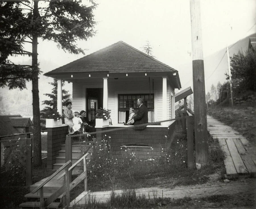 Group photo of the Samuels family on the porch of a house. A man and little boy are seated on the front of the porch and a little girl sits on a rocking horse next to two women.
