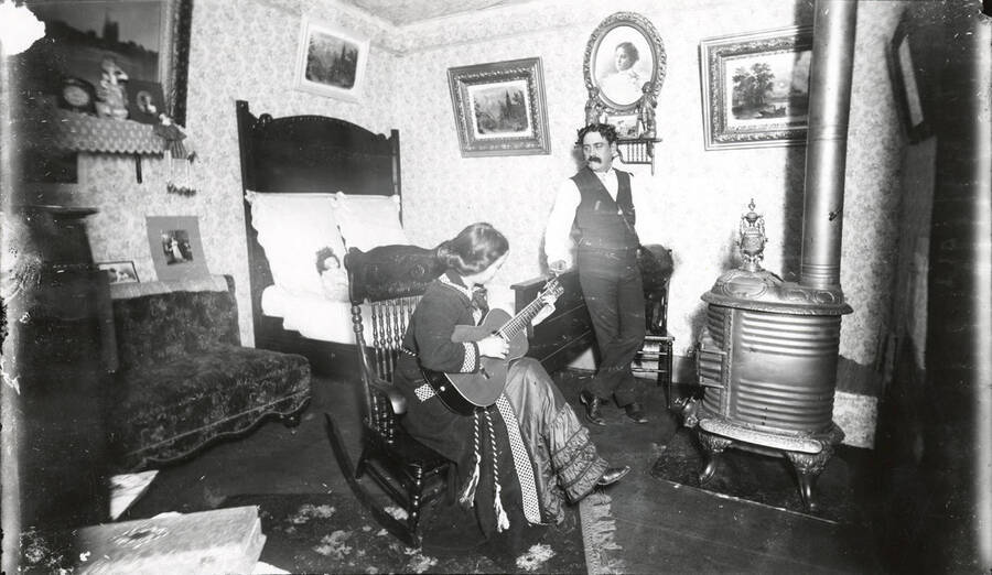Photograph of Mr. and Mrs. Hillard in the bedroom. Mrs. Hillard is playing guitar. Some degradation of the negative occurred prior to the printing of this photograph.