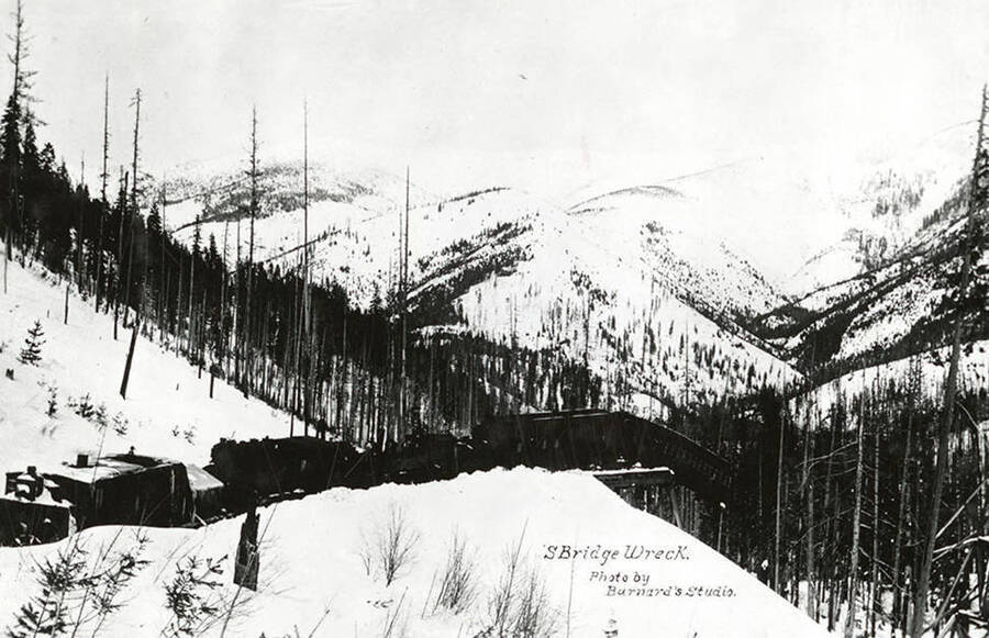 View of a train wreck at the S Bridge Trestle above Mullan, Idaho after a snow slide. Typed on the bottom: "S Bridge Wreck / Photo by / Barnard's Studio."