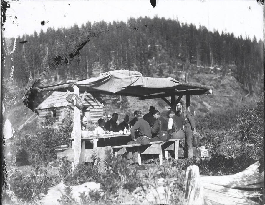Men eating at an outdoor table. The wing of a bird can be seen on the left hand side of the covered structure. The name "R.K. Stevenson" is written on the back of the photograph. Slight deterioration along the top left side of the negative prior to being printed.