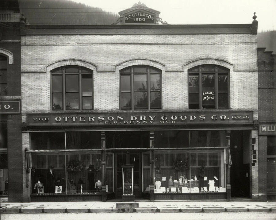 Exterior view of Otterson Dry Goods Company in Wallace, Idaho. Mannequins with dresses and other clothing displays can be seen through the windows of the store. Also shown is A.G. Kerns Law office.