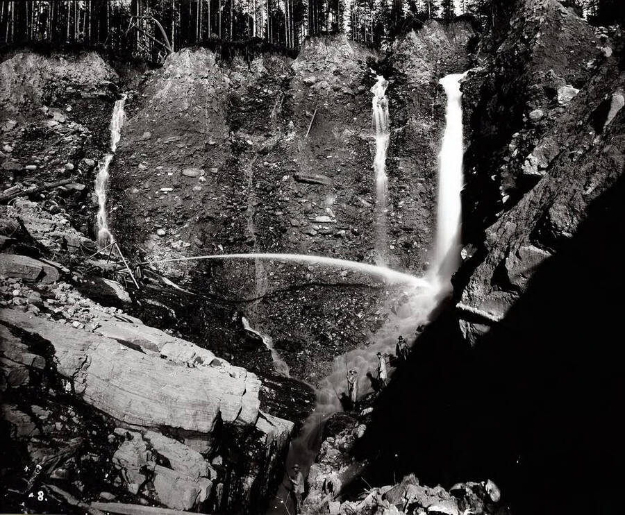Image shows hydraulic mining in Dream Gulch. Miners are seen holding a water hose and standing water fall and stream with mining tools.