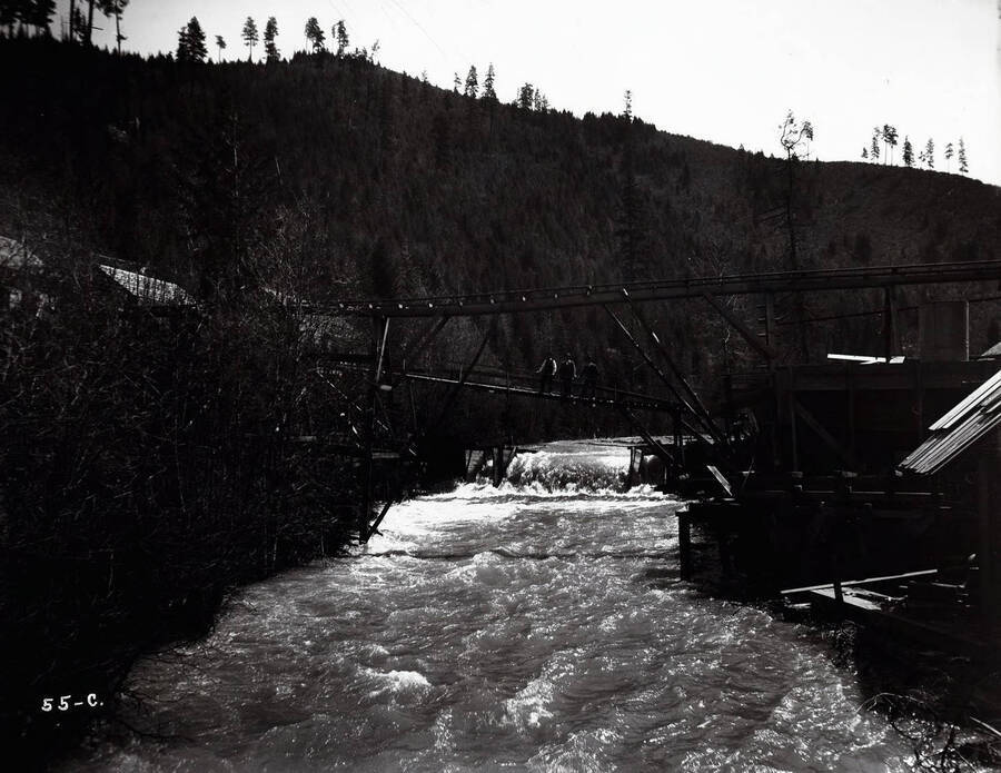 Image of the Coeur d'Alene Powder Co., in Wallace, Idaho; Three men are standing on bridge above the river.
