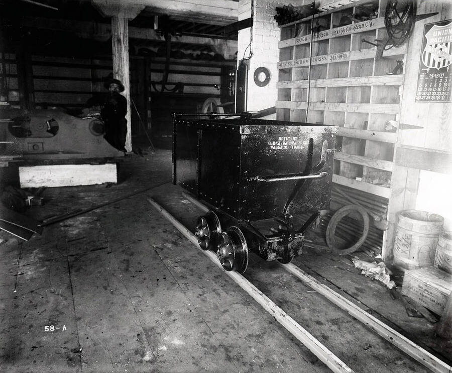 Image is of an ore car in the Coeur d'Alene Hardware Co., in Wallace, Idaho. An unidentified man crouches in the background.