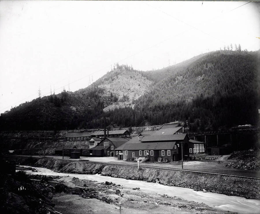 View of Hecla Mine in Burke, Idaho. Railroad cars sit in front of the buildings, with Canyon Creek running in front.