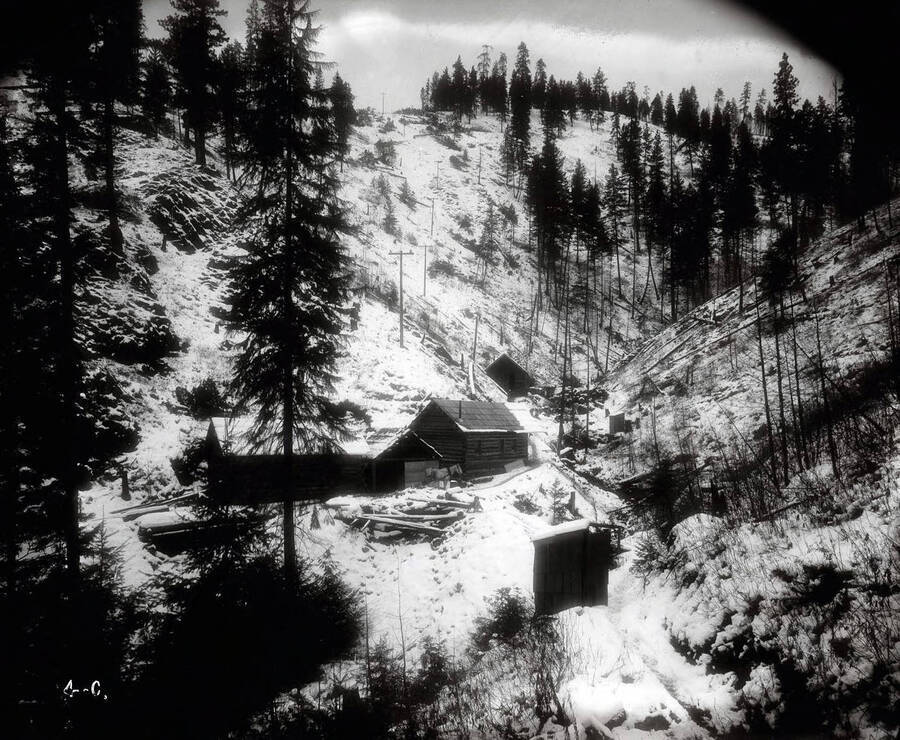 A snow covered setting shows a few buildings of Beebe Bros. Mine located in a gulch. A horse is pictured in front of one of the buildings.
