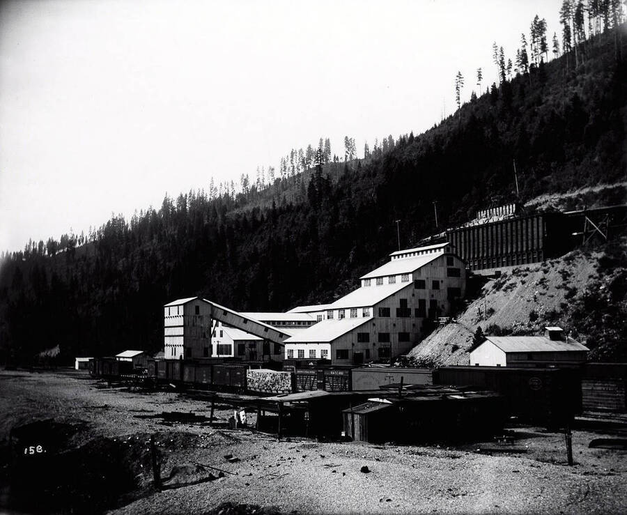 Image of the Hercules Concentrating Mill in Wallace, Idaho. Northern Pacific Railroad cars sit on the tracks in front of the buildings. One car is loaded with mine timbers.