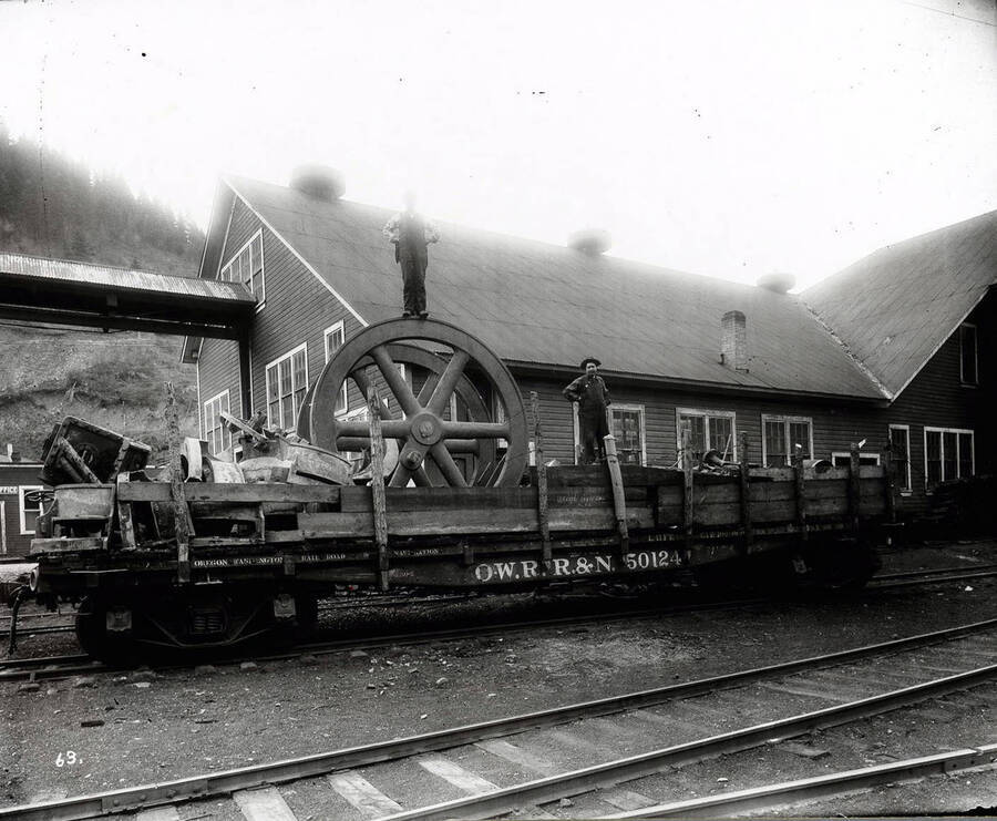 Image is of a rail car outside the Coeur d'Alene foundry. Two unidentified men are standing on the car with the letters O.W.R R&N 5012 [Oregon Washington Railroad and Navigation Company]