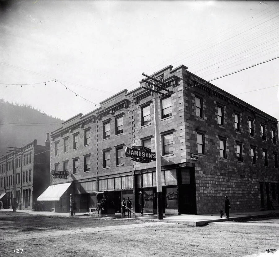 Exterior of Sweet's Hotel and William's and Fahle Jameson's Place. There is a shoe-shine stand and a constable talking to a man hosing off the sidewalk. Billiard Hall (Phister and Burns) is written on the two of the outside windows.