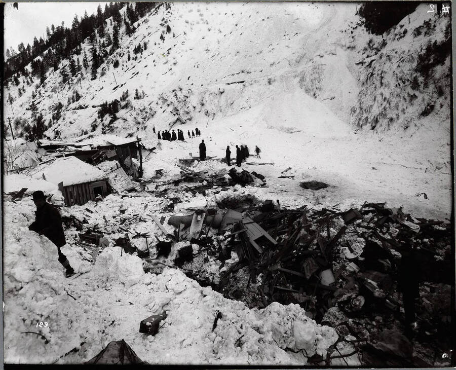 Image depicts building debris from a snow slide. Men, women and children standing at the scene.