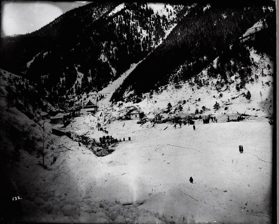 Far off view of a snow slide in Mace in East Shoshone County, near Burke, Idaho. Men and women are standing around at the base of the slide.