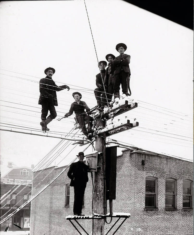 Image shows six telephone men in Wallace, Idaho standing on telephone pole. The  building is Masonic Temple (background) and Stevens Meat Market (Foreground).