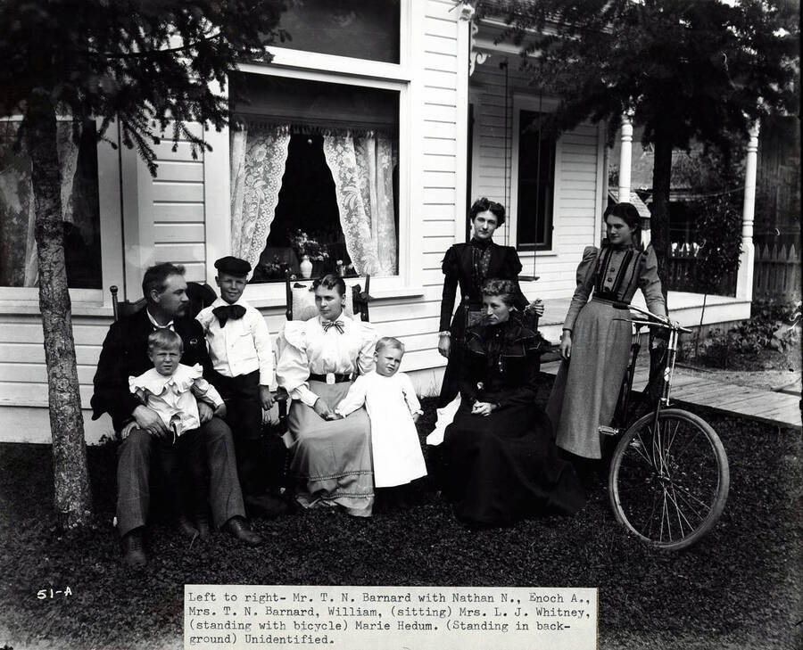 Image shows T.N. Barnard at his residence in Wallace, Idaho [1898] with a  group of people.  Mr. Barnard was a photographer and the owner of Barnard's Studio in Wallace, Idaho.  Caption below image: "Left to right - Mr. T. N. Barnard with Nathan N., Enoch A., Mrs. T.N. Barnard,  William, (sitting) Mrs. L.J. Whitney, (standing with bicycle) Marie Hedum. (Standing in background) Unidentified."