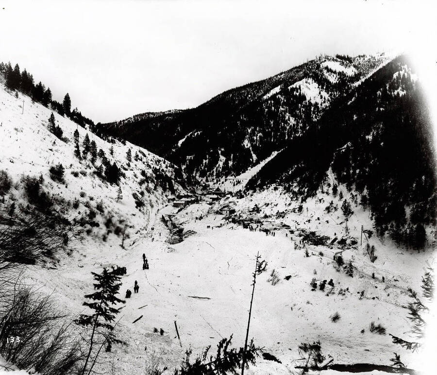 Far off view of a snow slide in Mace in East Shoshone County, near Burke, Idaho. Men and women are standing around at the base of the slide.