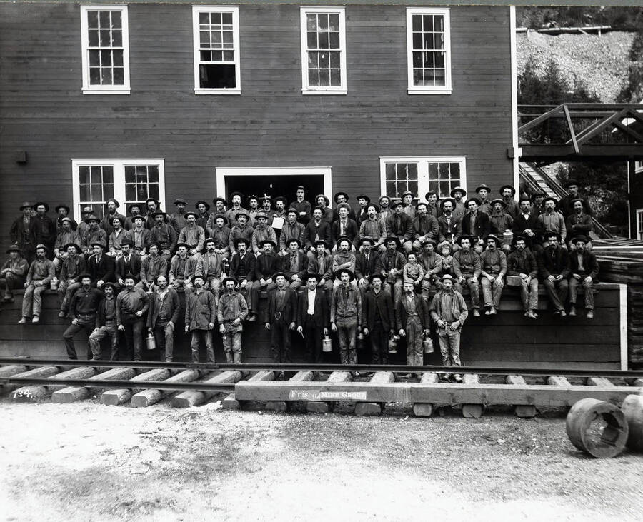 Large group of men standing and sitting in front of a wooden building in with railroad tracks. Many are holding lunch pails. Caption on front: "Frisco Mine Group."