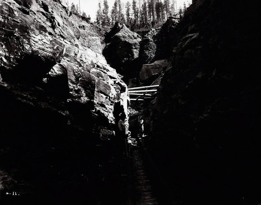 Image is of the Arizona Placer mine (hydraulic mining) located in  Murray, Idaho; In the image is a sluice box and riffles. A couple of miners are also present.