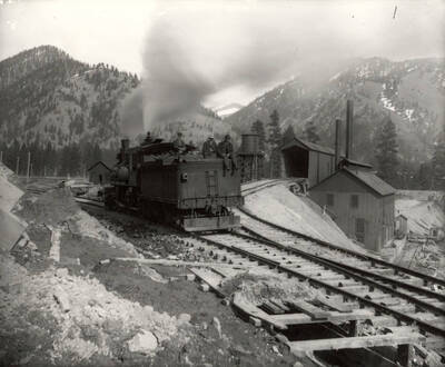 Men sitting on top of a train engine on the railroad tracks, with a water storage tank in the background. Taken at the Amador Mine in Cedar Creek, Montana.