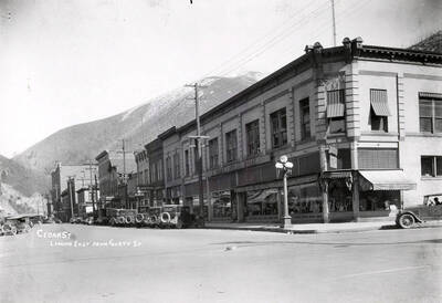 Caption reads: "Cedar St. Looking east from Fourth Street." Note this could actually be Fifth Street in Wallace, Idaho. Numerous buildings and businesses line the street, which has cars parked on it."