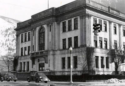 Exterior view of the Shoshone County Courthouse in Wallace, Idaho. Two cars are parked out front and hills can be seen in the distance.