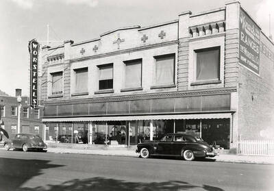Exterior view of the Worstell Company Building in Wallace, Idaho. Cars are parked out front and furniture can be seen in the windows of the building.