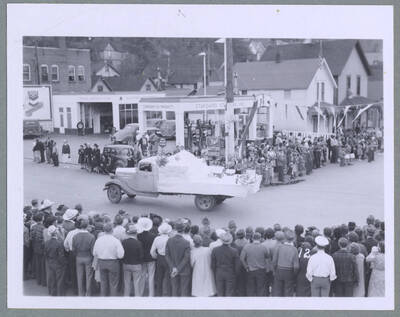Women marching with flags in front of a car towing a float with a model home on it during the Elks Roundup Parade in Wallace, Idaho.