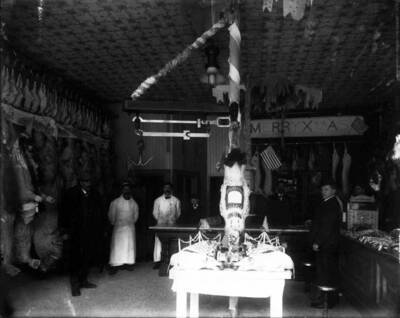 Whole beef and carcasses of chickens can be seen hanging along the wall of Stevens Meat Market. Note sign that reads "merry Xmas", and two whole roasted pigs sitting on a table.