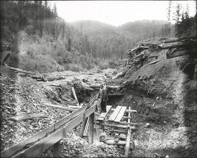 North side, Coeur d'Alene Mining District (Murray area). A man stands next to the flume in the photograph.