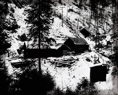 A snow covered setting shows a few buildings of Beebe Bros. Mine located in a gulch. A horse is pictured in front of one of the buildings.